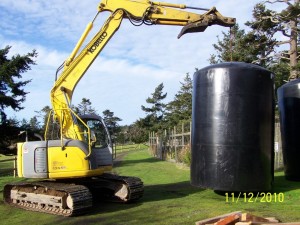 Excavator lifting large black tank outdoors.